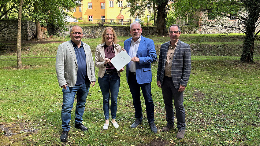 Ein Frau und drei Männer stehen in einem Park, im Hintergrund ist ein Schloss zu sehen. Die Frau und einer der Männer halten zusammen ein Dokument in Händen.