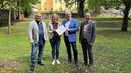 Ein Frau und drei Männer stehen in einem Park, im Hintergrund ist ein Schloss zu sehen. Die Frau und einer der Männer halten zusammen ein Dokument in Händen.