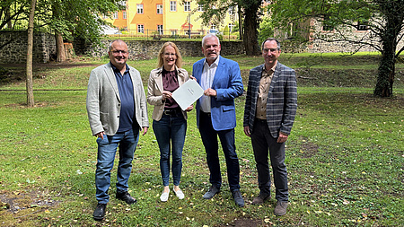 Ein Frau und drei Männer stehen in einem Park, im Hintergrund ist ein Schloss zu sehen. Die Frau und einer der Männer halten zusammen ein Dokument in Händen.
