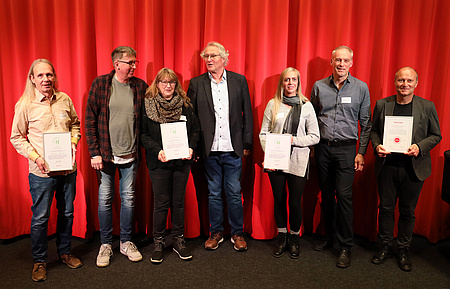 Sieben Personen stehen auf einer Bühne, einige halten Urkunden in die Kamera.