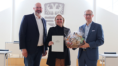 Zwei Männer und eine Frau stehen nebeneinander. Sie halten eine Urkunde und einen Geschenkkorb in den Händen