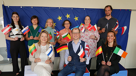 Eine große Gruppe von Menschen sitzt und steht vor einer Europa-Flagge, die an der Wand hängt.