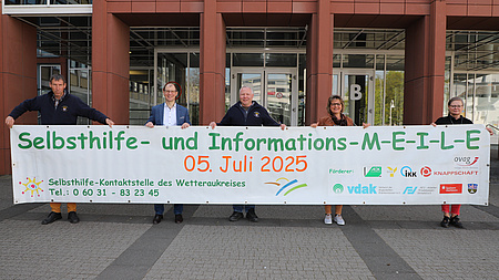 Fünf Leute vor einem Gebäude. Sie halten gemeinsam ein großes Banner, auf dem "Selbsthilfemeile" steht.