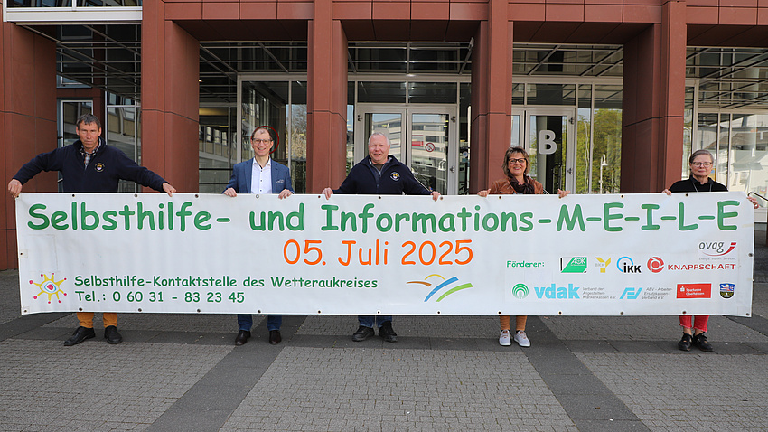 Fünf Leute vor einem Gebäude. Sie halten gemeinsam ein großes Banner, auf dem "Selbsthilfemeile" steht.