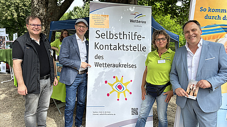 Drei Männer und eine Frau stehen um ein Banner herum und lächeln. Darauf steht "Selbsthilfe-Kontaktstelle des Wetteraukreises".