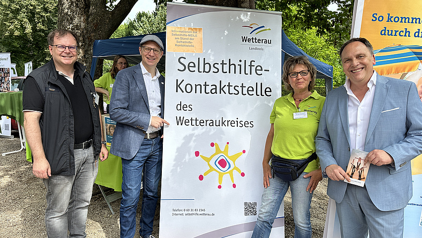 Drei Männer und eine Frau stehen um ein Banner herum und lächeln. Darauf steht "Selbsthilfe-Kontaktstelle des Wetteraukreises".