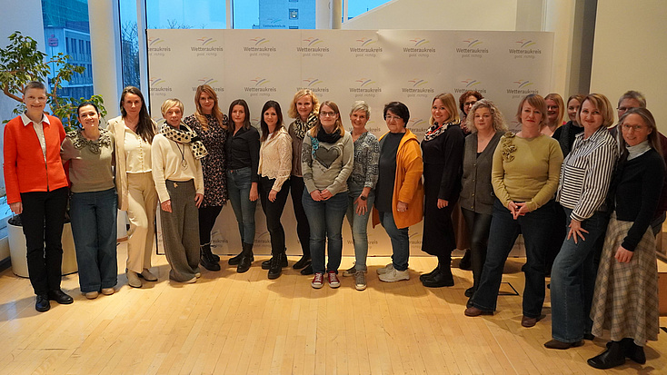 Eine Gruppe von etwa zwei Dutzend Frauen steht in einem Saal vor einer Fensterfront und einer Fotowand.