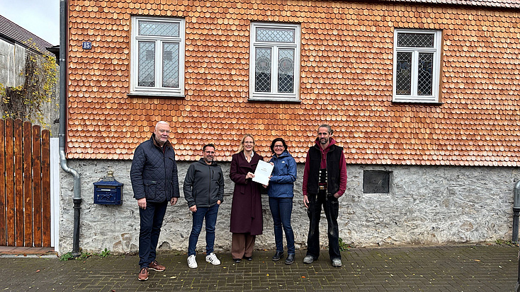 Zwei Frauen und drei Männer stehen auf einem Bürgersteig vor einer mit Holzschindeln verkleideten Fassade eines Hauses. Die beiden Frauen halten gemeinsam ein Dokument in Händen.