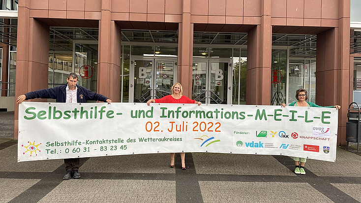 Von links: Dr. Reinhold Merbs, Erste Kreisbeigeordnete und Sozialdezernentin Stephanie Becker-Bösch und Anette Obleser. Alle halten ein 6 m langes Banner, welches auf die Selbsthilfe-MEILE hinweist.