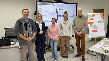 Fünf Personen stehen vor einer digitalen Tafel.