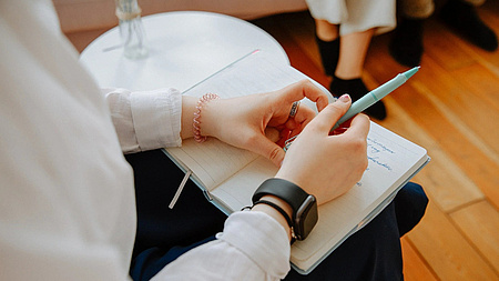 Eine Person sitzt auf einem Stuhl neben einem Tisch und hält einen Stift und ein Notizbuch im Schoß. Ihr gegenüber sitzen weitere Personen.