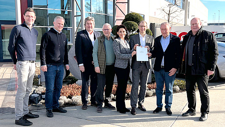 Mehrere Männer und eine Frau stehen nebeneinander. Drei von ihnen halten gemeinsam ein Schriftstück. Im Hintergrund ein Gebäude und Autos.
