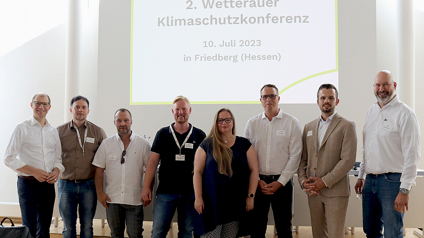 Sieben Männer und eine Frau stehen nebeneinander. Im Hintergrund steht auf der Leinwand: 2. Wetterauer Klimaschutzkonferenz