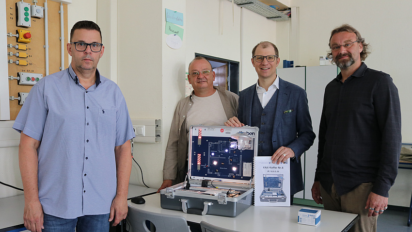 Vier Männer stehen in einem Klassenzimmer rund um einen Experimentierkoffer.