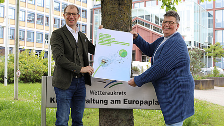 Ein Mann und eine Frau stehen vor einem Gebäude. Sie halten ein Plakat in die Luft. Darauf steht "Herzlich willkommen bei Ihrer Schwerbehindertenvertretung."