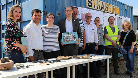 Neun Leute stehen vor einem Baucontainer. Im Vordergrund auf einem Tisch sieht man antike Scherben.