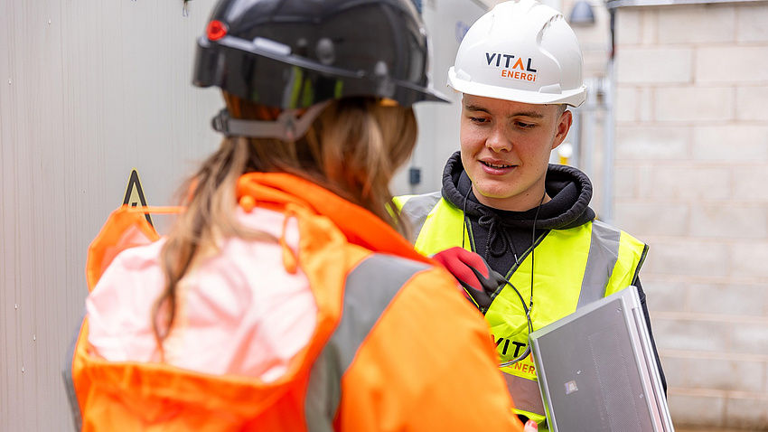 Eine Frau und ein Mann in neonfarbener Warn- und Arbeitskleidung und mit Schutzhelmen unterhalten sich auf einer Baustelle.