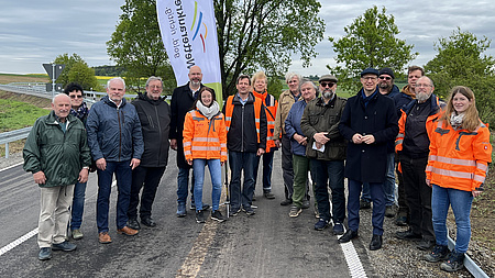 Eine Gruppe von Leuten steht auf einer sanierten Straße. Einige Personen tragen bunte Warnwesten.