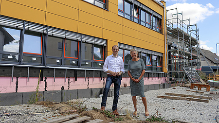 Ein Mann und eine Frau stehen auf einer Baustelle, im Hintergrund ist ein fast fertiger Neubau mit gelber Außenverkleidung zu sehen.
