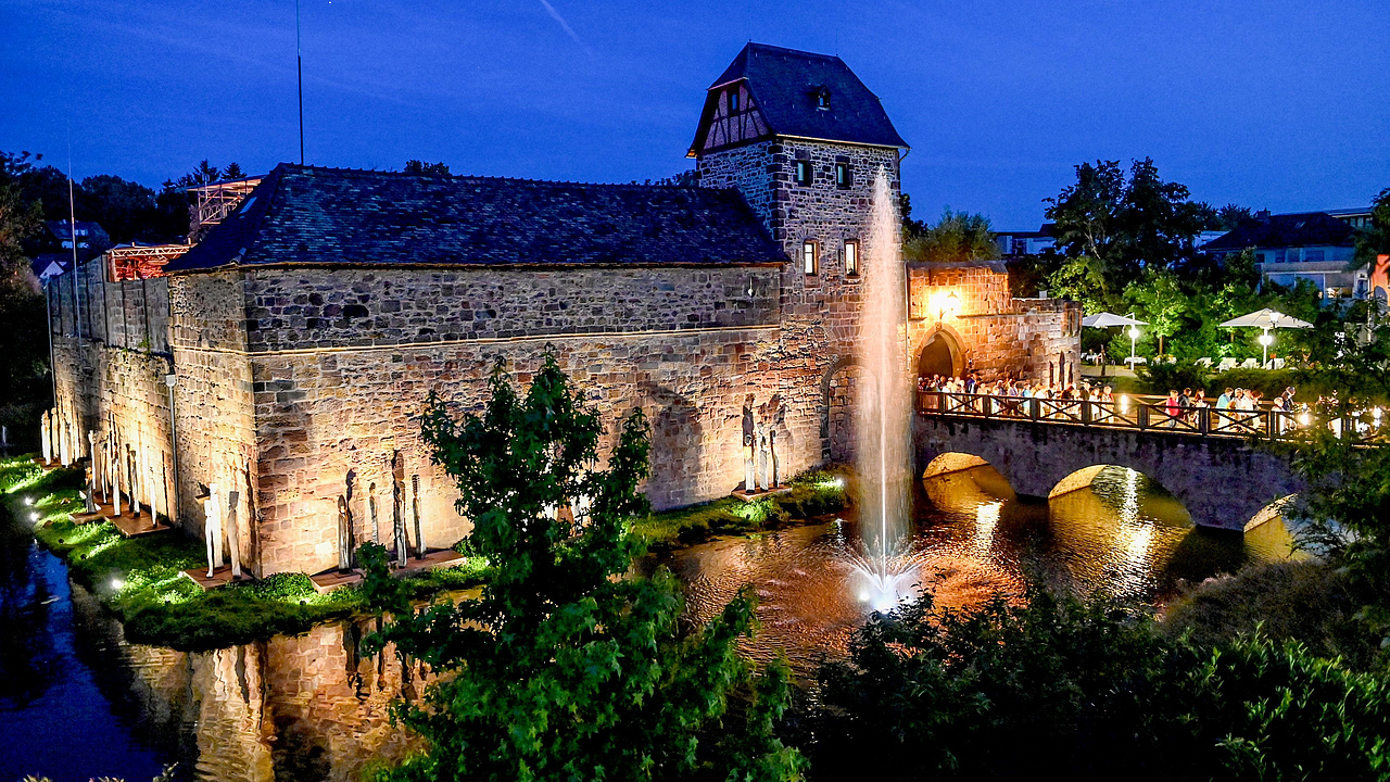 Eine mittelalterliche Vilbeler Wasserburg bei Sonnenuntergang. Vor der beleuchteten Burg ist einen Burggraben voll mit Wasser und einem Brunnen. Eine Brücke voll mit Menschen verbindet die Burg mit dem Festland. 
