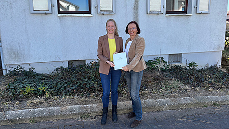 Zwei Frauen stehen in einem Hof vor einer Häuserfassade und halten gemeinsam ein Dokument in Händen.