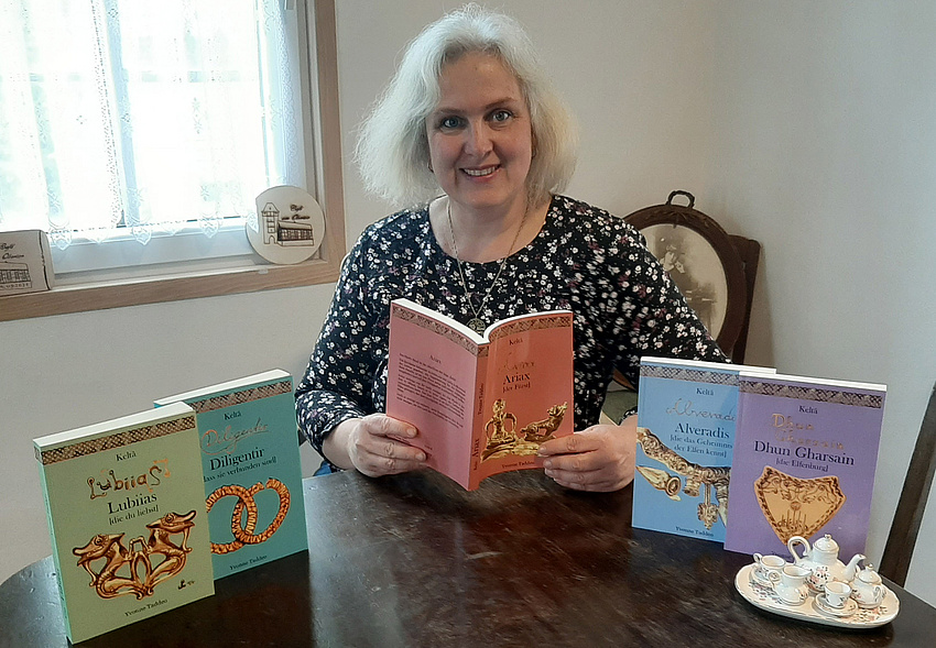 Eine Frau sitzt an einem runden Tisch, lächelt in die Kamera und hält ein Buch in der Hand. Links und rechts sind jeweils zwei Bücher zu sehen.