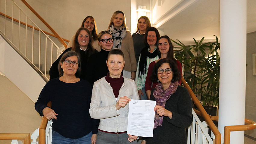 Zehn Frauen in einem Treppenhaus, zwei von ihnen halten einen Förderbescheid in die Kamera.