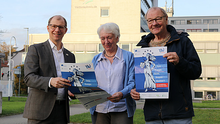 Drei Personen stehen im Freien nebeneinander und präsentieren den Flyer für den Europäischen Wettbewerb 2026.