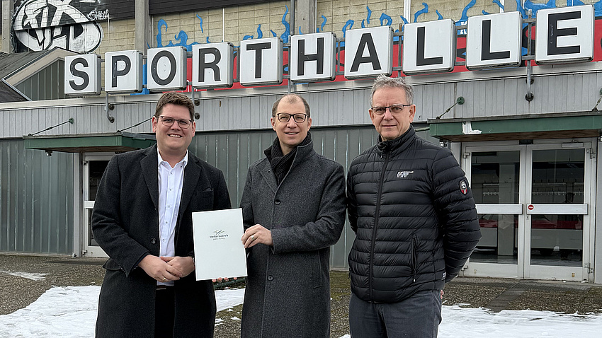 Drei Männer stehen vor dem Eingang einer großen Halle, auf der die Aufschrift Sporthalle steht. Einer der Männer übergibt einem zweiten ein Dokument.
