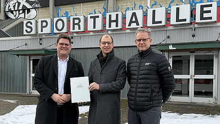 Drei Männer stehen vor dem Eingang einer großen Halle, auf der die Aufschrift Sporthalle steht. Einer der Männer übergibt einem zweiten ein Dokument.