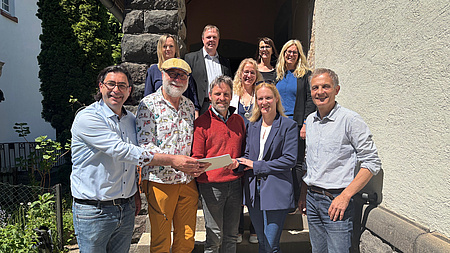 Mehrere Frauen und Männer stehen vor dem Eingang eines Hauses. Drei der Männer und eine der Frauen in der ersten Reihe halten gemeinsam ein Dokument in Händen.