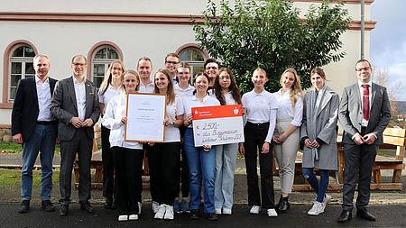 Eine Gruppe Jugendlicher und Erwachsener steht vor einem Schulgebäude. Einige haben ein großformatiges Schriftstück in der Hand.