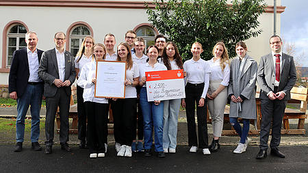 Eine Gruppe von Personen steht im Freien vor einem Baum. Sie halten eine Urkunde und einen Scheck mit der Aufschrift "Wetterauer Schulpreis" in den Händen.