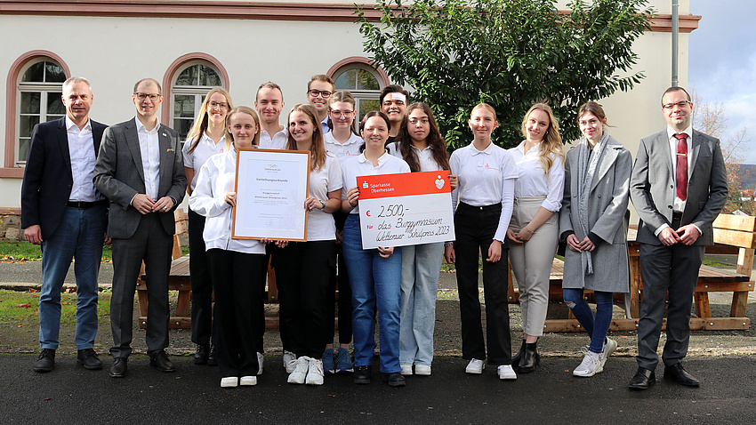 Eine Gruppe von Personen steht im Freien vor einem Baum. Sie halten eine Urkunde und einen Scheck mit der Aufschrift "Wetterauer Schulpreis" in den Händen.
