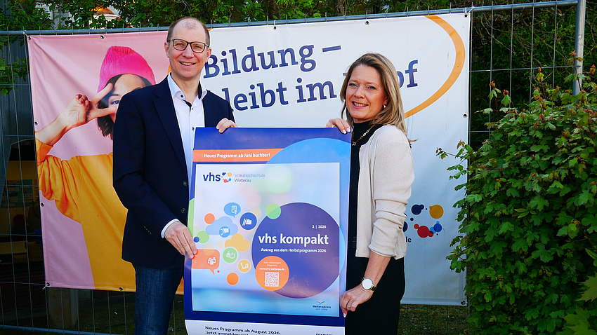 Eine Frau und ein Mann stehen im Freien. Hinter ihnen ist ein Werbebanner aufgehängt, und beide halten gemeinsam ein Plakat hoch.