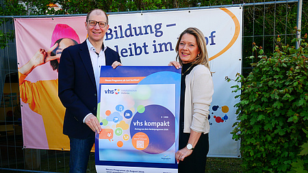 Eine Frau und ein Mann stehen im Freien. Hinter ihnen ist ein Werbebanner aufgehängt, und beide halten gemeinsam ein Plakat hoch.