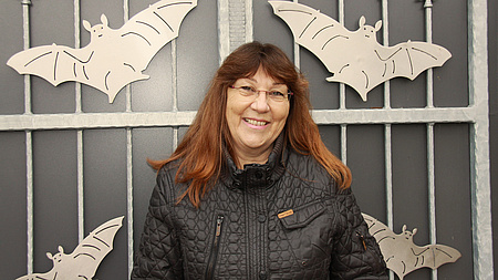 Eine Frau mit Brille und rotbraunen Haaren steht vor einem Metalltor, auf dem Fledermäuse abgebildet sind.