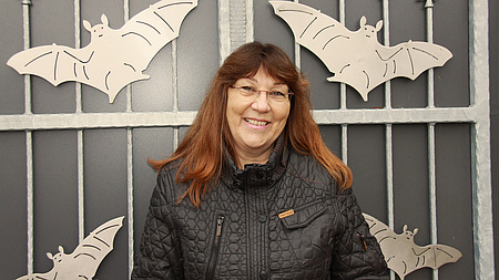 Eine Frau mit Brille und rotbraunen Haaren steht vor einem Metalltor, auf dem Fledermäuse abgebildet sind.