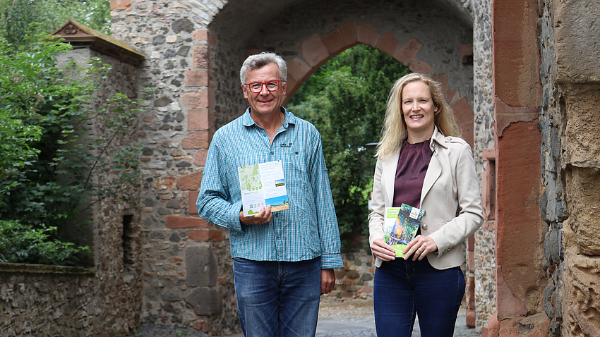 Zwei Menschen stehen in einem alten Torbogen und halten zwei Broschüren in der Hand.