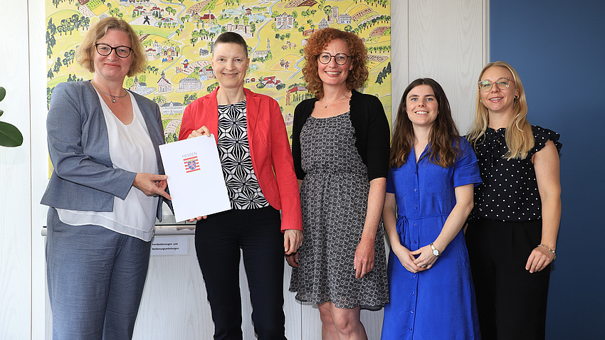 Fünf Frauen stehen in einem Raum vor einem Gemälde. Zwei Frauen der Gruppe halten gemeinsam eine Dokumentenmappe in Händen.