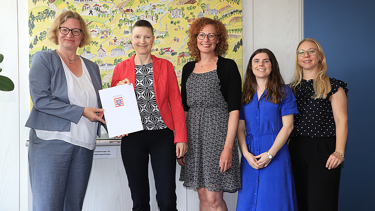Fünf Frauen stehen in einem Raum vor einem Gemälde. Zwei Frauen der Gruppe halten gemeinsam eine Dokumentenmappe in Händen.