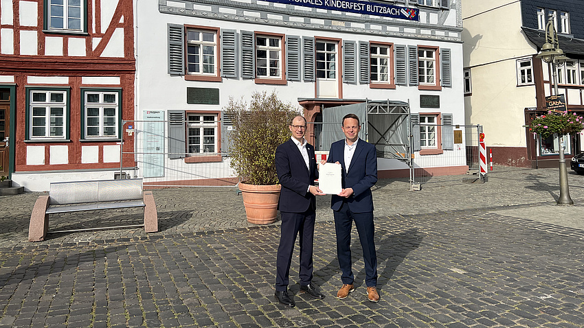 Landrat Jan Weckler und Bürgermeister Sascha Huber stehen vor einem Fachwerkhaus.