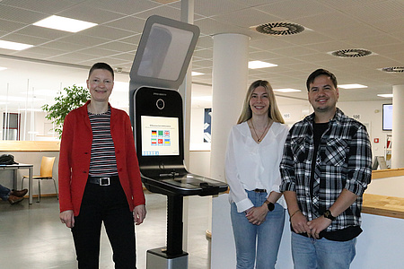 Zwei Frauen und ein Mann stehen in einem großen Büroraum vor einem technischen Gerät, mit dem man Passfotos anfertigen kann.