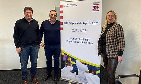 Eine Frau und zwei Männer stehen neben einem Rollup. Auf diesem steht, dass der Regionalverband Rhein-Main der Johanniter-Unfallhilfe beim Katastrophenschutzpreis 2021 den 2. Platz gewonnen hat.
