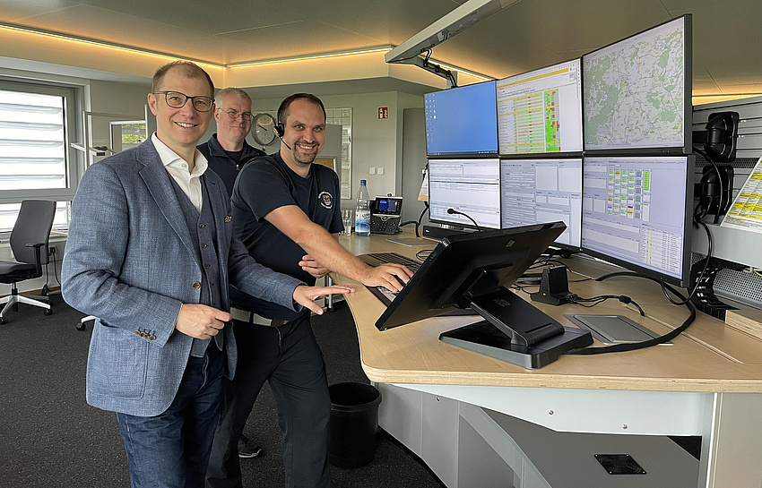 Drei Männer stehen an einem höhenverstellbaren Schreibtisch. Auf diesem stehen mehrere Bildschirme über- und nebeneinander.