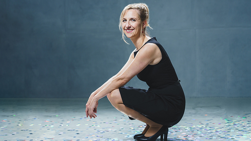 Eine Frau in kurzem schwarzen Kleid sitzt in der Hocke und lächelt in die Kamera