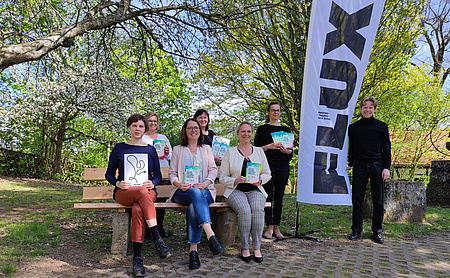 Drei Frauen sitzen auf eienr Parkbank, drei Frauen stehen dahinter. Alle haben ein Prospekt in der Hand. Rechts steht ein Mann, hinter ihm ein Banner mit dem Schriftzug Flux.