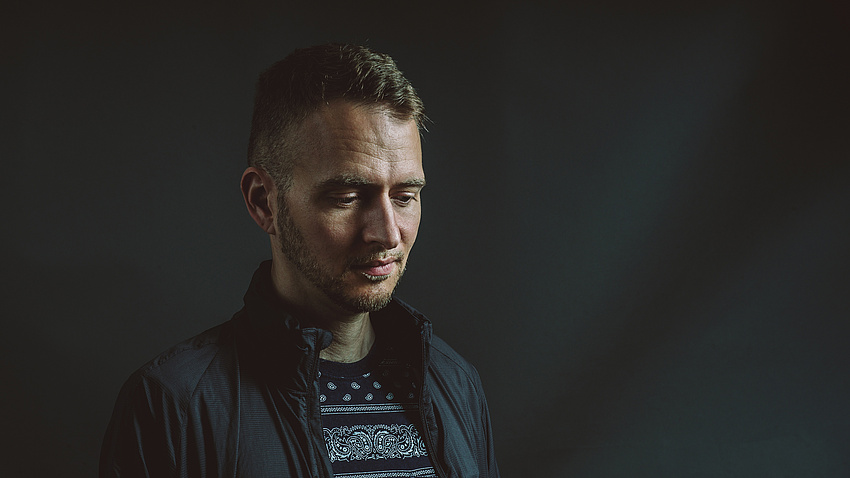 Ein Mann blickt nachdenklich nach unten, er steht vor einer schwarzen Wand.