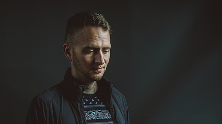 Ein Mann blickt nachdenklich nach unten, er steht vor einer schwarzen Wand.