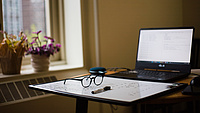 Das Bild zeig einen aufgeklappten Laptop auf einem Tisch. Davor liegen eine Brille und ein Stift.
